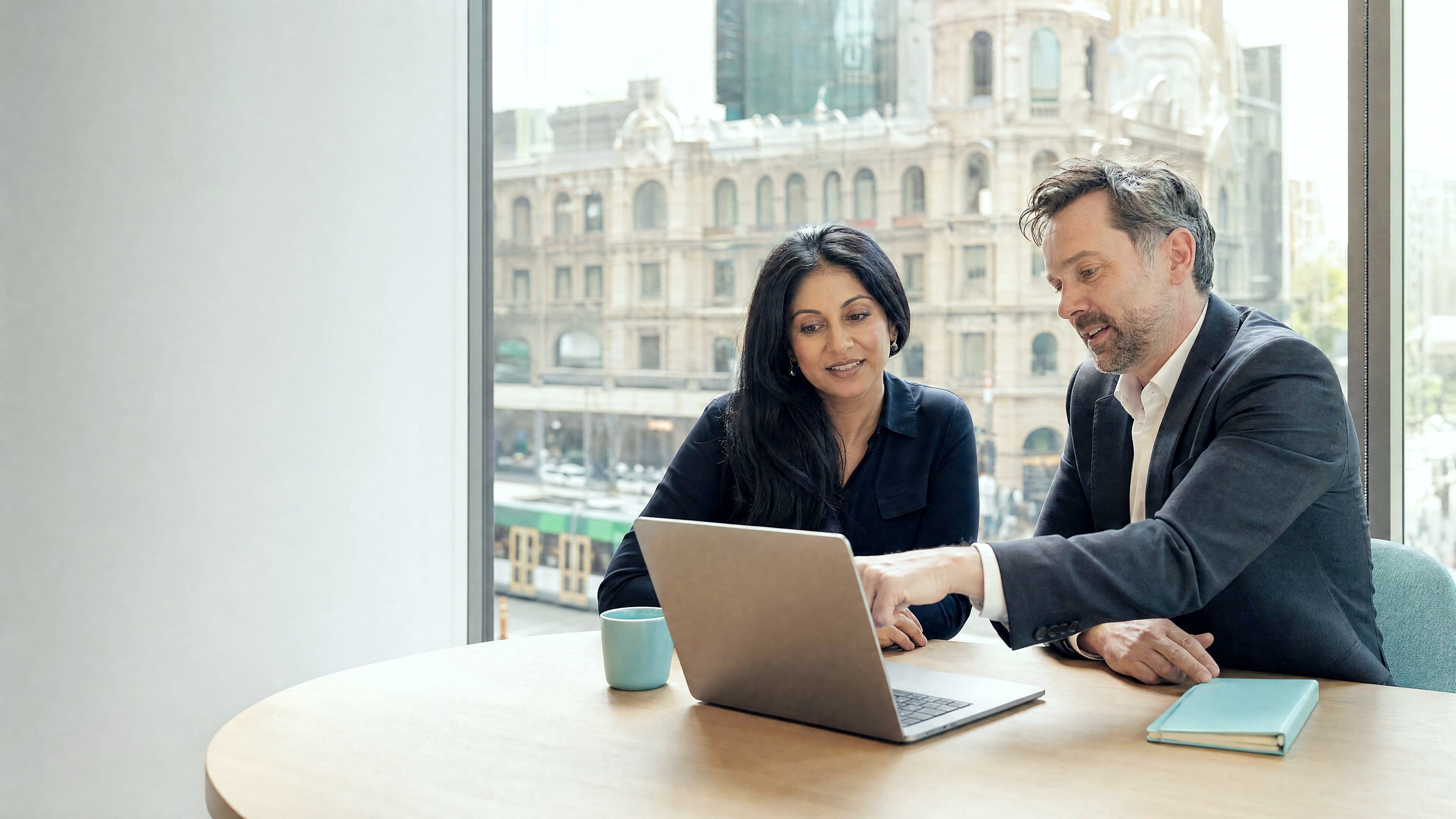 Two professionals reviewing a laptop together at a timber table in an office above Collins Street.